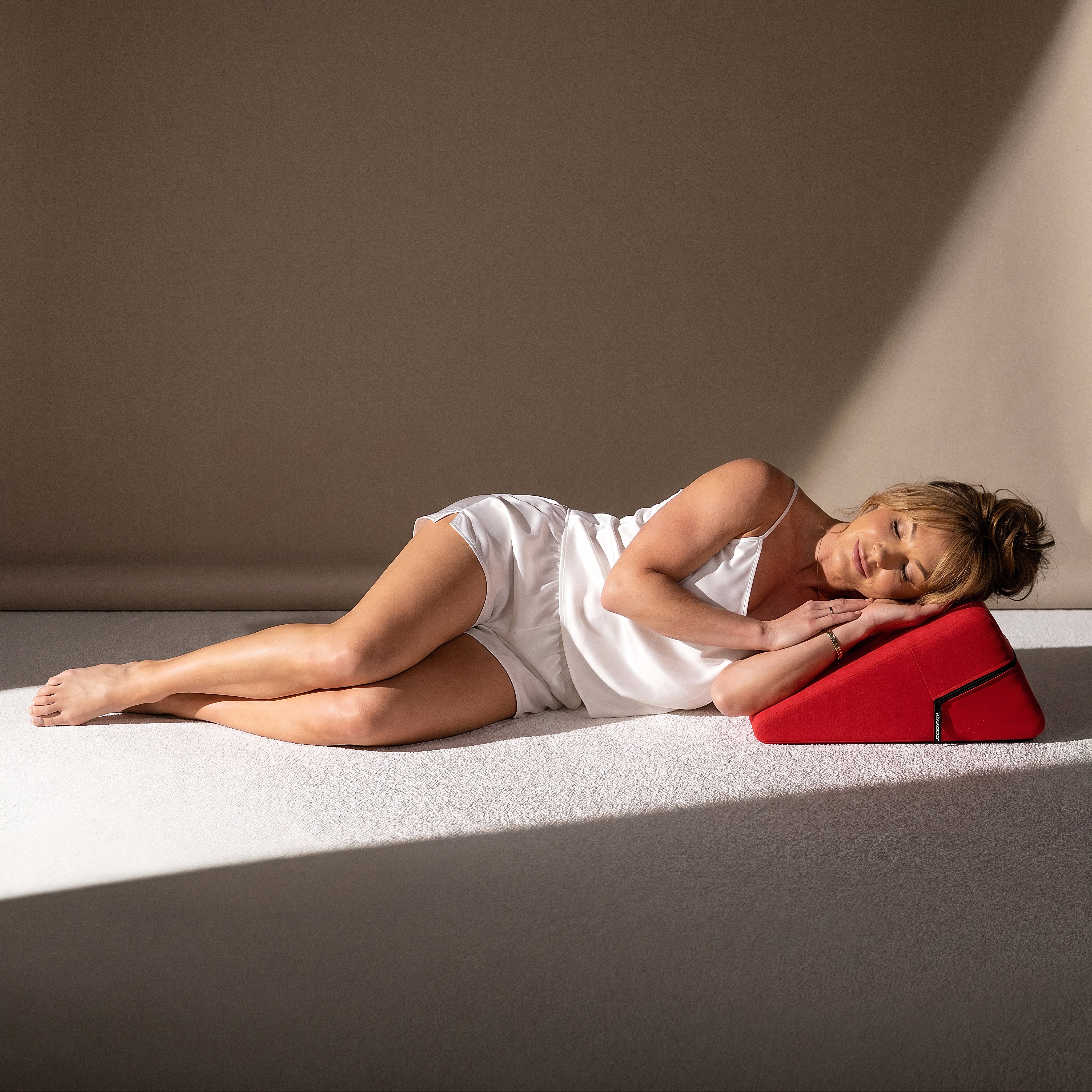 Woman in white satin sleepwear resting her head on the red Liberator Wedge in a sunlit studio