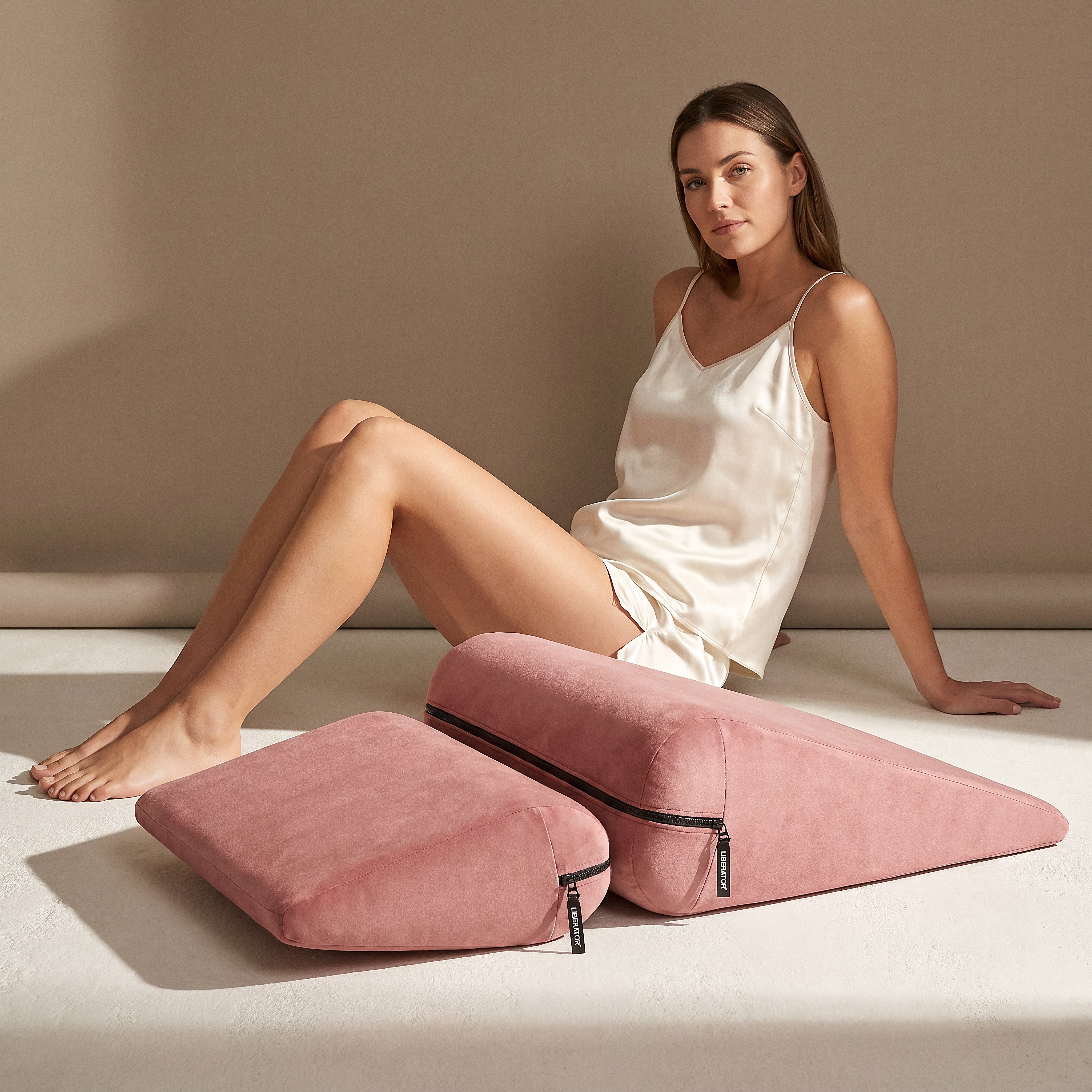 Woman in ivory satin sleepwear seated beside the rose Liberator Jaz Duet cushions in a sunlit studio