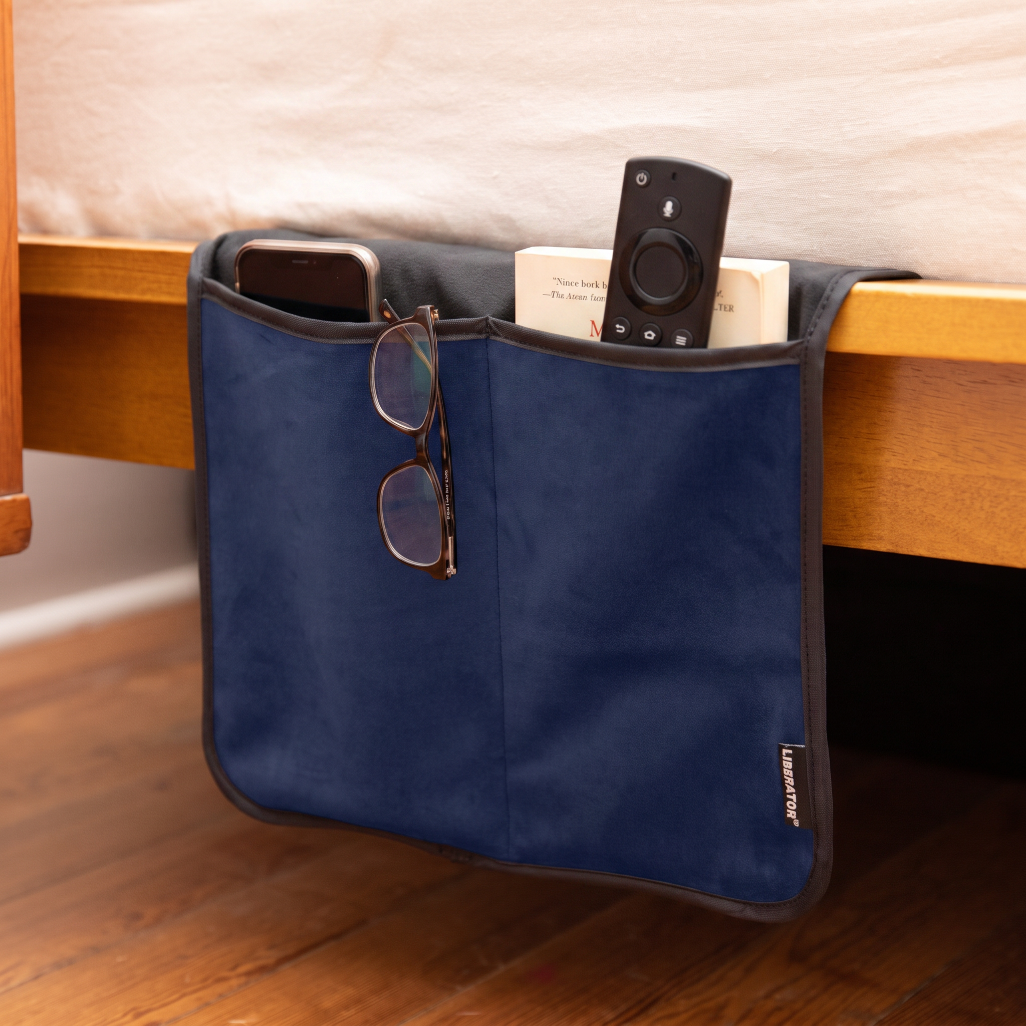 Fascinator Bedside Caddy in navy hanging on the bed, holding a phone, reading glasses, remote and a book